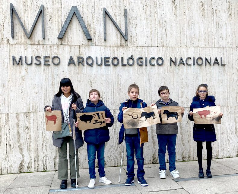 Un grupo de niñas y niños, bajo las corpóreas del museo Arqueológico Nacional. En sus manos sujetan cada uno un papel de tipo Kraft en el que hay dibujada una pintura rupestre que representa a un animal prehistórico